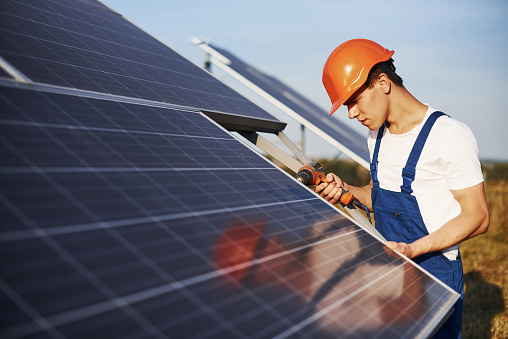 Using cordless screwdriver. Male worker in blue uniform outdoors with solar batteries at sunny day.